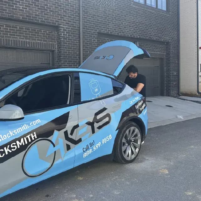 A technician in a black shirt leans into the raised rear hatch of a light blue wrapped 4Keys Locksmith service vehicle parked in a paved alley beside a brick building with garage doors